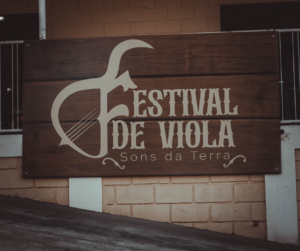 Placa de madeira com o logo do Festival de Viola – Sons da Terra, exibindo uma ilustração estilizada de viola caipira ao lado do nome do evento.