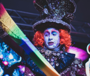 Artista caracterizado como o Chapeleiro Maluco, com maquiagem colorida, peruca laranja e figurino detalhado com botões e brilhos, segurando um tecido colorido durante apresentação do projeto Feira ao Ar Livro.