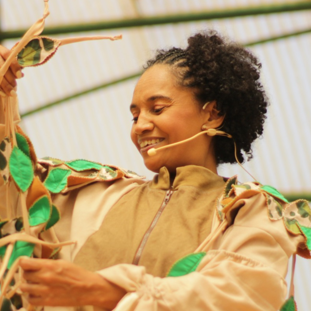 Jussara, uma mulher sorrindo durante uma apresentação, usando figurino bege decorado com folhas e microfone de cabeça, segurando elementos cênicos em um ambiente iluminado.