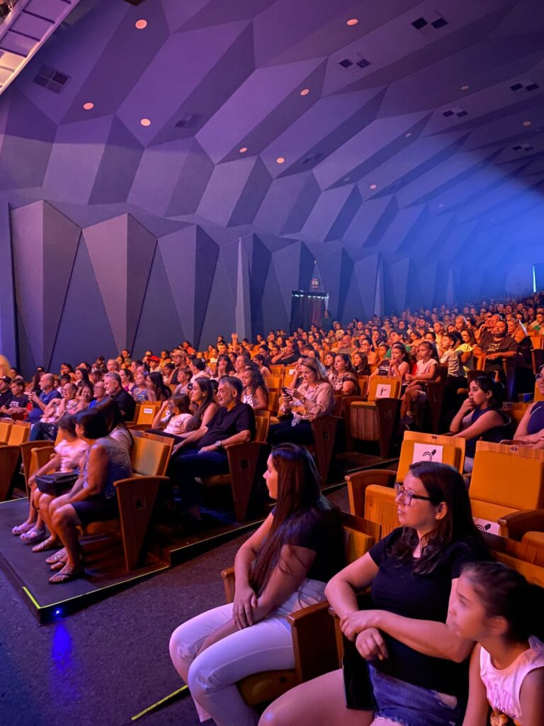 Público assistindo espetáculo cultural em teatro durante apresentação com grande participação